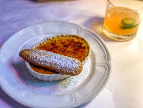 A creamy dessert in a dish with a cookie, dusted with powdered sugar, and a cocktail in the background.