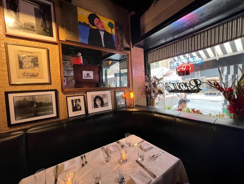 Cozy restaurant corner with a table set for two, framed art on the walls, and a view of the street outside.