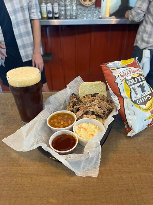 A plate of pulled pork with coleslaw, baked beans, barbecue sauce, a bun, chips, and a pint of dark beer on a table.