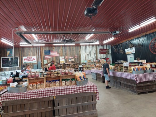 Interior of a rustic market with wooden stalls, checkered tablecloths, and various food products. An American flag hangs above.