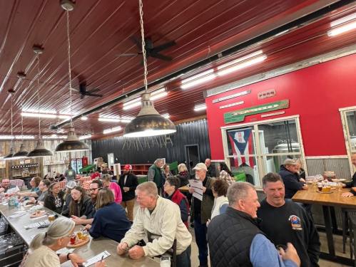 A lively bar scene with many patrons enjoying drinks and conversation in a rustic setting with red walls and metal accents.