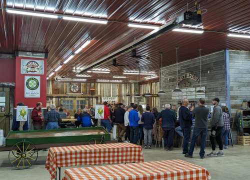 A crowd of people gathered inside a rustic venue with wooden decor, enjoying a social event. Tables with checkered cloths are visible.
