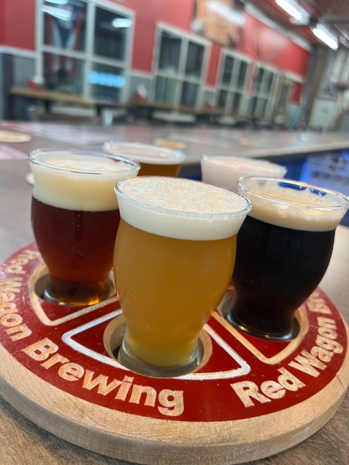 A wooden flight tray holds six glasses of different craft beers, showcasing various colors and foam levels.