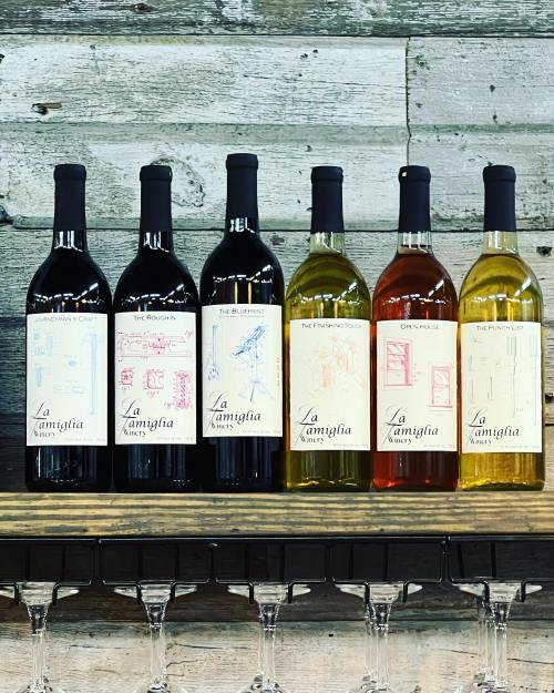 A selection of six wine bottles on a wooden shelf, featuring various labels and colors, with wine glasses below.