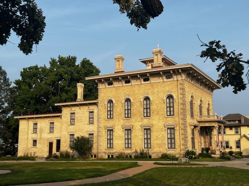 Historic yellow mansion with ornate architecture, large windows, and a well-maintained lawn under a clear blue sky.