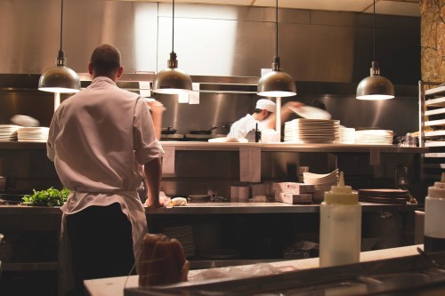 Two chefs work in a busy kitchen, preparing dishes and plating food under warm overhead lights.