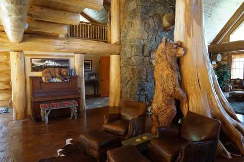 Cozy log cabin interior featuring a carved bear, wooden pillars, leather chairs, and a piano against a stone wall.