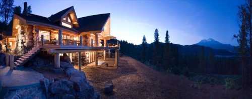 A panoramic view of a mountain cabin at dusk, surrounded by trees and mountains in the background.