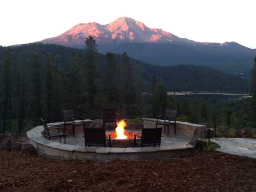 A fire pit surrounded by chairs, with a mountain backdrop and trees at sunset.