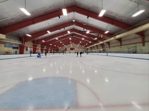 A spacious indoor ice rink with skaters gliding on the ice under a high ceiling and red beams.