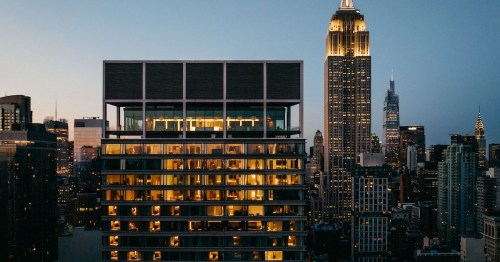 A modern building with illuminated windows in the foreground, with the Empire State Building lit up in the background at dusk.