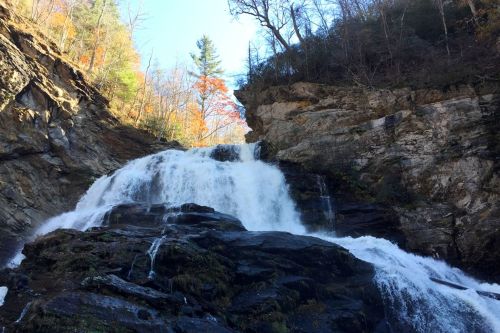 A cascading waterfall flows over rocky cliffs, surrounded by trees with autumn foliage.