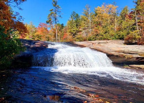 A serene waterfall cascades over rocks, surrounded by vibrant autumn foliage under a clear blue sky.