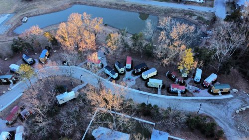 Aerial view of a winding road lined with colorful tiny homes near a pond, surrounded by trees in a rural setting.
