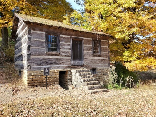 A rustic log cabin surrounded by vibrant autumn foliage and a stone foundation, with a sign nearby.