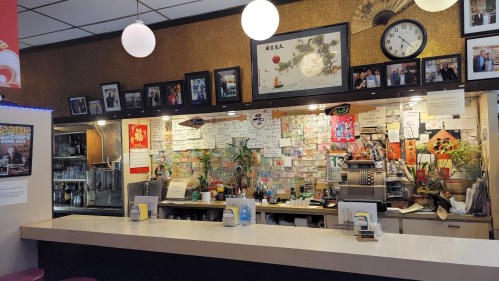 A cozy restaurant counter with a variety of photos, notes, and decorations on the wall, featuring plants and a clock.