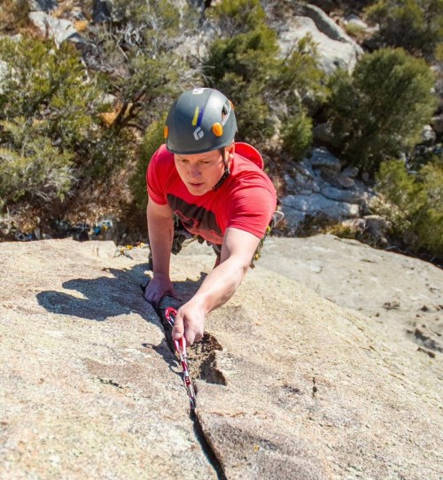 A climber in a helmet reaches for a handhold on a rocky surface, surrounded by trees and boulders.