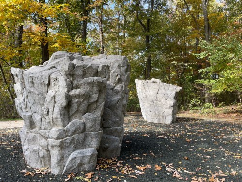 Two large, textured rock sculptures in a wooded area with autumn foliage and fallen leaves on the ground.