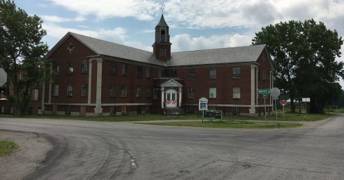 A large red brick building with a clock tower, situated at a road intersection, surrounded by trees and grassy areas.