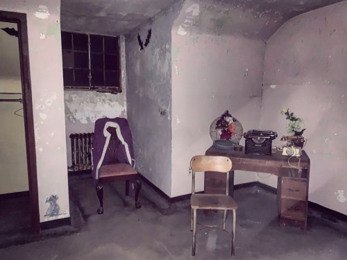 An empty, dimly lit room with peeling walls, a desk with a typewriter, and a chair beside a window.