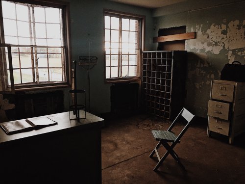 An empty, dimly lit room with two windows, a chair, a desk, and peeling paint on the walls.