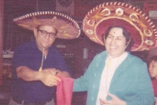 Two people wearing sombreros smile and shake hands, with a festive background and a child in the corner.