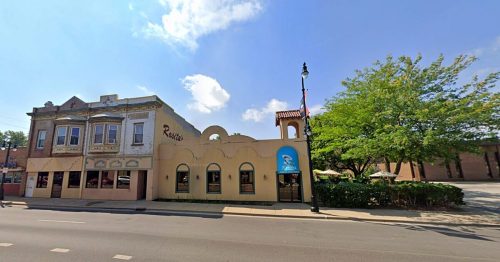 A colorful building with arched windows and a sign, next to a historic structure, under a clear blue sky.
