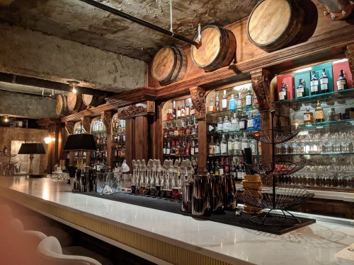 A stylish bar with a white countertop, shelves of various spirits, and wooden barrels on the wall.
