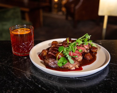 A plate of steak with roasted vegetables and sauce, accompanied by a cocktail in a glass.