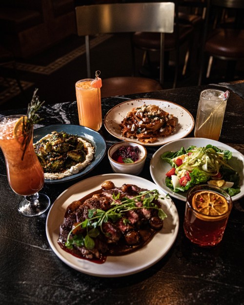 A table spread with various dishes: grilled meat, salad, hummus, and colorful cocktails in a cozy setting.