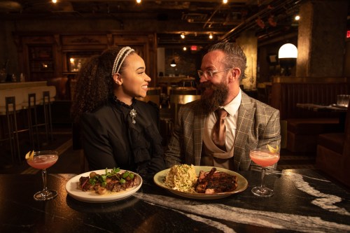 A couple enjoys a romantic dinner at a restaurant, smiling at each other with plates of food and drinks on the table.