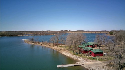 A serene lake view with a red cabin, trees, and a dock under a clear blue sky.