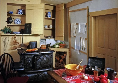A vintage kitchen with a cast-iron stove, wooden shelves, a table with a yellow bowl, and various kitchen items.