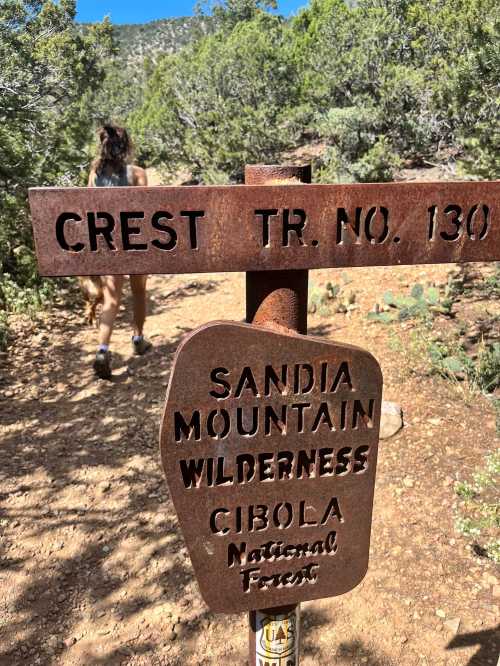 A rusted trail sign for Crest Trail No. 130 in Sandia Mountain Wilderness, Cibola National Forest, with a hiker in the background.