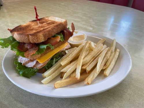 A stacked sandwich with bacon, turkey, cheese, and lettuce, served with a side of fries and dipping sauce.