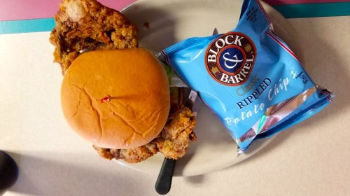 A plate with fried chicken, a sandwich bun, and a bag of Block & Barrel rippled potato chips.