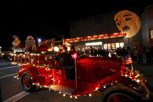 A festive parade featuring a vintage fire truck adorned with lights and Santa, with a large face projected on a building.