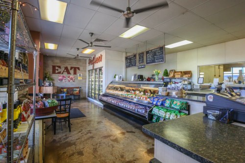 Interior of a deli with a display case, shelves of snacks, and a cozy seating area. "EAT" sign on the wall.