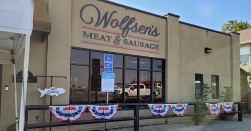 Exterior of Wolfson's Meat & Sausage with patriotic bunting and a sign, featuring large windows and a parking area.