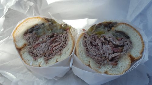 A close-up of a sandwich cut in half, filled with shredded meat and pickles, wrapped in white paper.