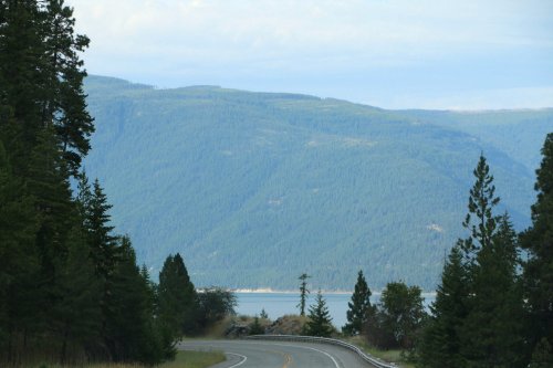 A winding road leads towards a serene lake, surrounded by lush green mountains and trees under a cloudy sky.