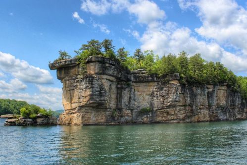 A rocky cliff with trees atop, rising from a calm lake under a blue sky with fluffy clouds.