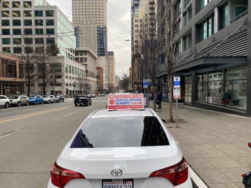 A white car with a sign reading "Instruction Car" parked on a city street surrounded by tall buildings.