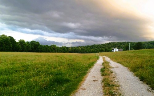 A gravel path leads through a grassy field towards a small white building under a cloudy sky.