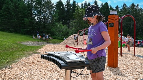 A child plays a xylophone at a playground, surrounded by greenery and other children enjoying the outdoors.