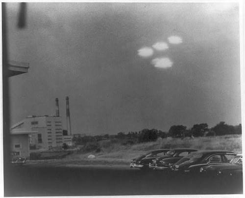 Black and white photo of cars parked near a factory, with unidentified flying objects in the sky above.
