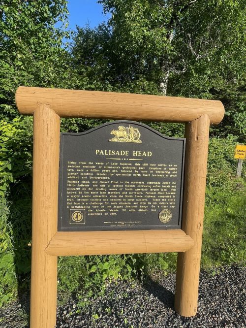 A wooden sign titled "Palisade Head" with information about the area, surrounded by greenery and blue sky.