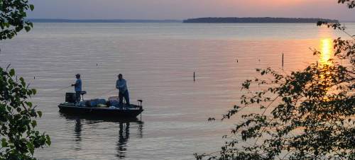 Two fishermen on a boat in calm waters at sunset, surrounded by trees and a serene landscape.