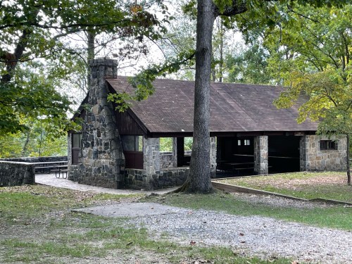 A stone picnic shelter surrounded by trees in a park, with a gravel path leading to it.