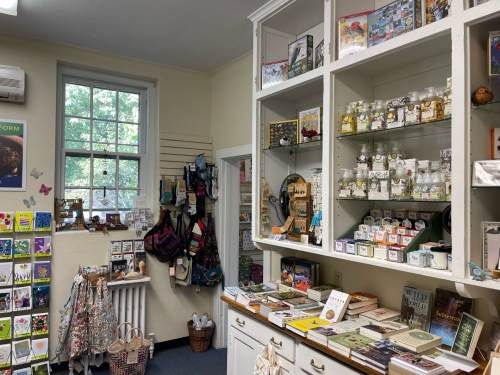 A cozy shop interior featuring shelves of books, gifts, and various decorative items, with a window letting in natural light.